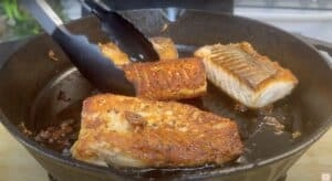 Sizzling salmon fillets in a cast iron pan, being flipped with tongs for perfect sear.